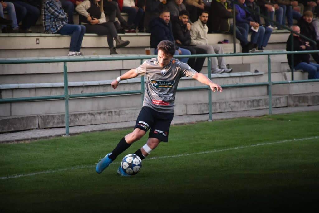 Fußballspieler beim Dribbling am Spielfeldrand, Zuschauer auf der Tribüne beobachten das Spiel aufmerksam.