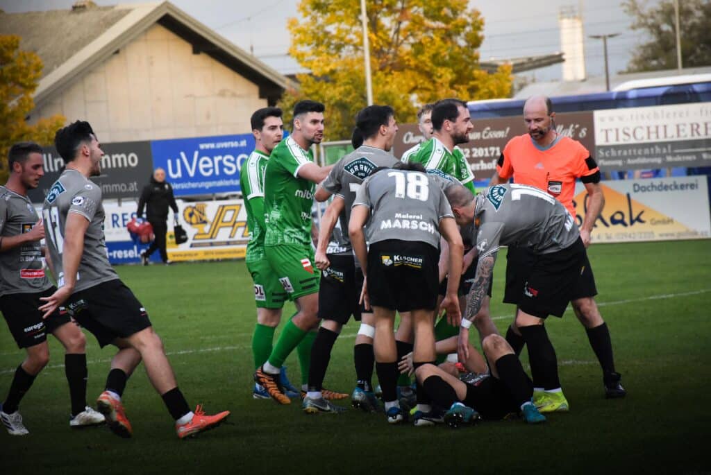 Fußballspieler diskutieren auf dem Feld nach einem Zweikampf, während der Schiedsrichter die Situation beobachtet.
