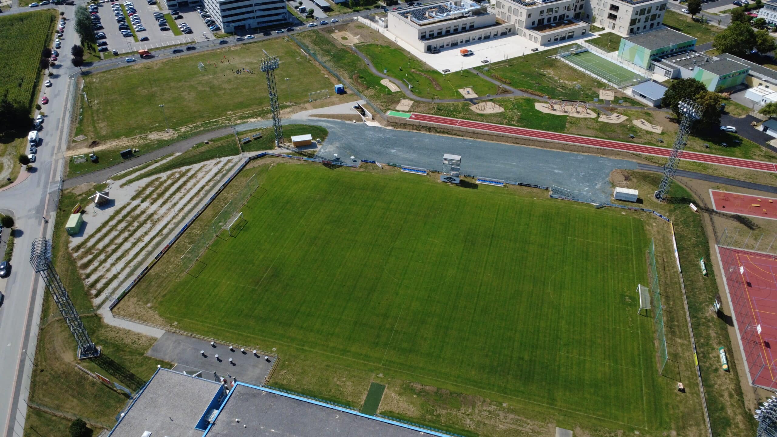 Luftaufnahme eines Fußballplatzes mit umliegenden Sportanlagen und Parkplätzen, grüne Rasenfläche im Mittelpunkt.