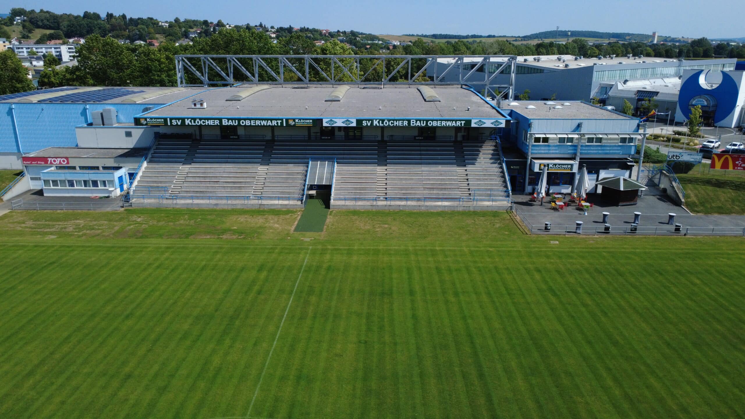 Luftaufnahme eines leeren Fußballstadions mit grüner Spielfläche, Sitzplätzen und blauer Tribüne in Oberwart.