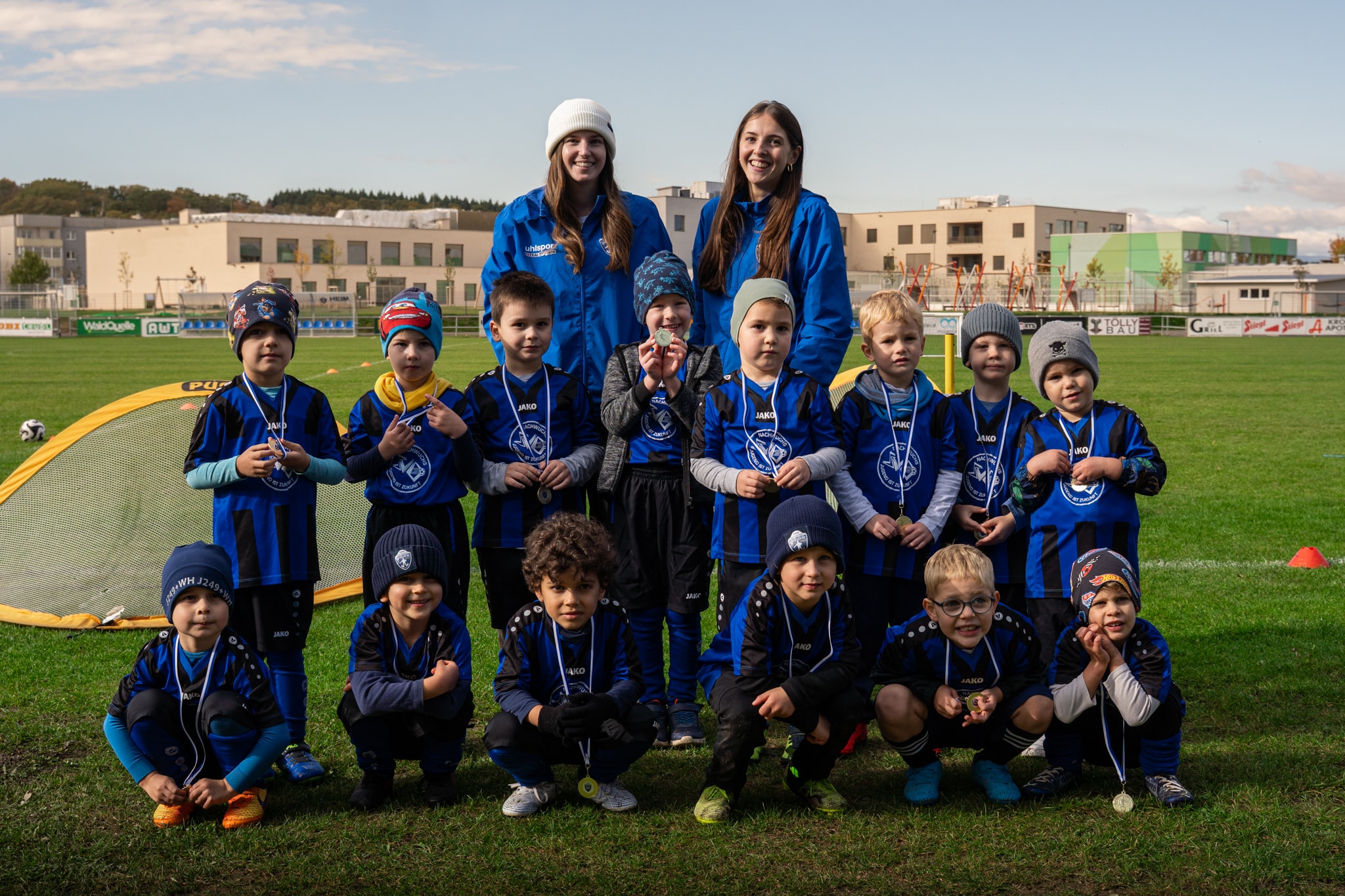 Junge Fußballmannschaft mit Trainerinnen im blauen Trikot posiert nach einem erfolgreichen Spiel auf dem Sportplatz.