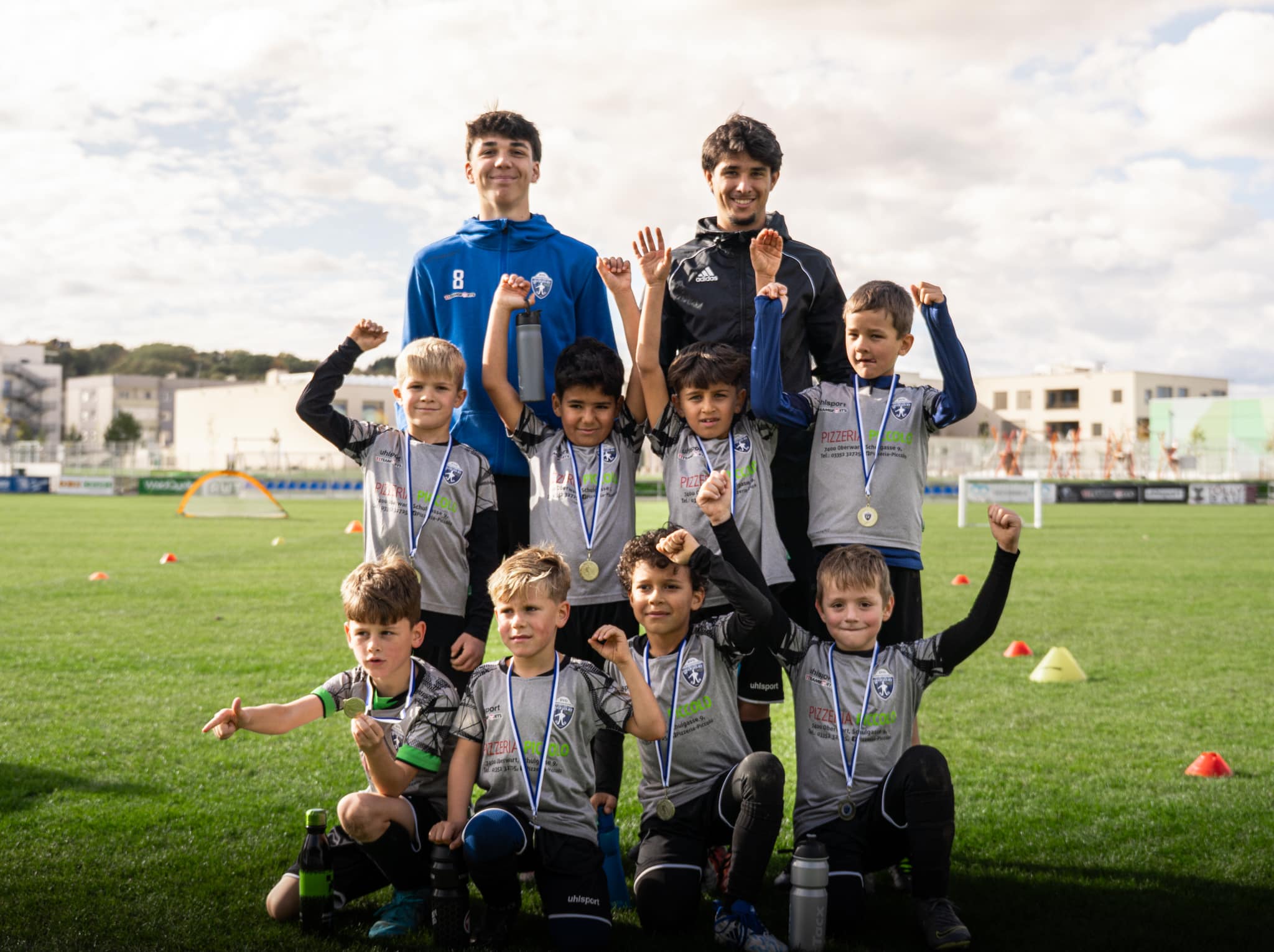 Kinder-Fußballteam feiert mit Medaillen auf dem Spielfeld an einem sonnigen Tag.