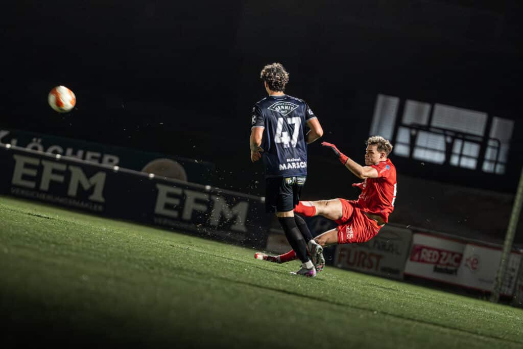 Fußballspiel bei Nacht: Spieler im Zweikampf auf dem Rasen, dynamische Action und fliegender Ball im Flutlicht.