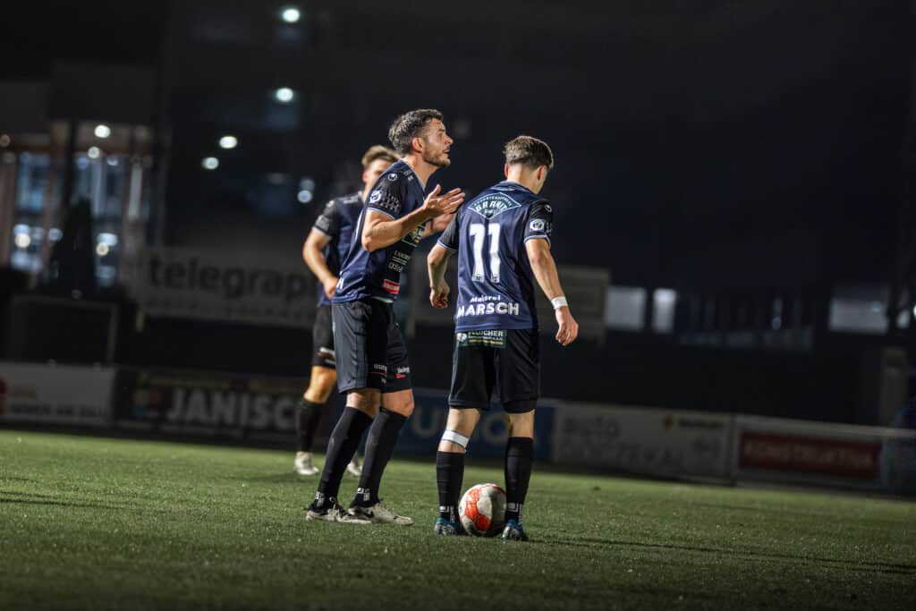 Fußballspieler diskutieren auf dem Spielfeld bei Nachtspiel, Fokus auf Mannschaftsstimmung und Strategie.