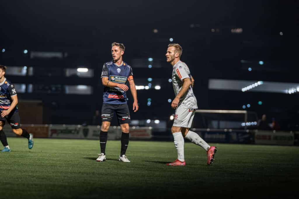 Zwei Fußballspieler im Spiel, blaues und graues Trikot, beleuchtetes Stadion bei Nacht, Spielspannung.