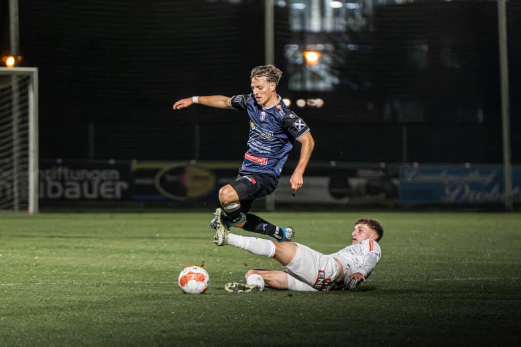 Fußballspieler wird während eines Spiels bei einem Tackling angegriffen, Ball im Vordergrund, intensives Spiel auf dem Feld.