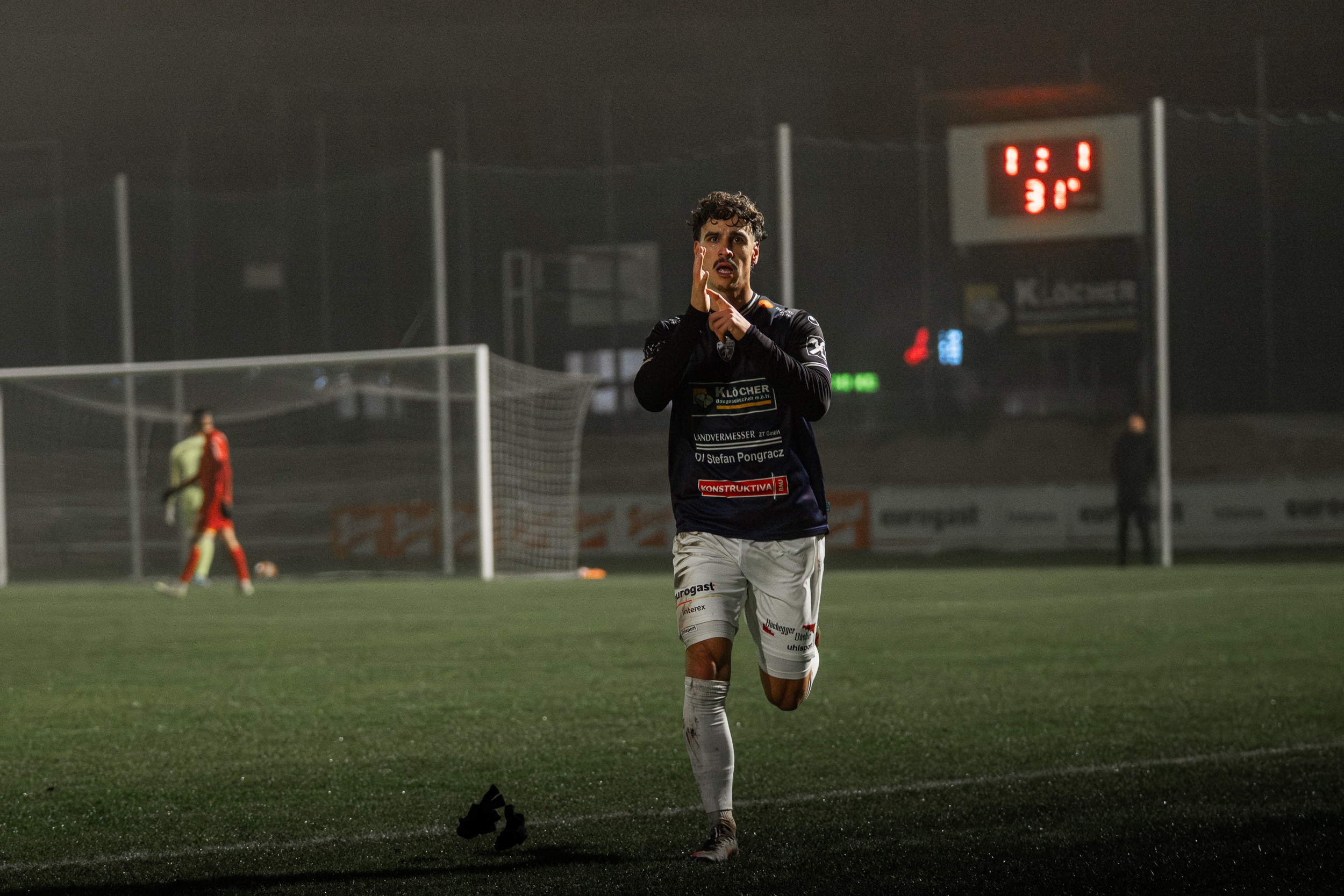 Fußballspieler jubelt auf dem Spielfeld nach einem Tor, Anzeigetafel zeigt Spielstand an.