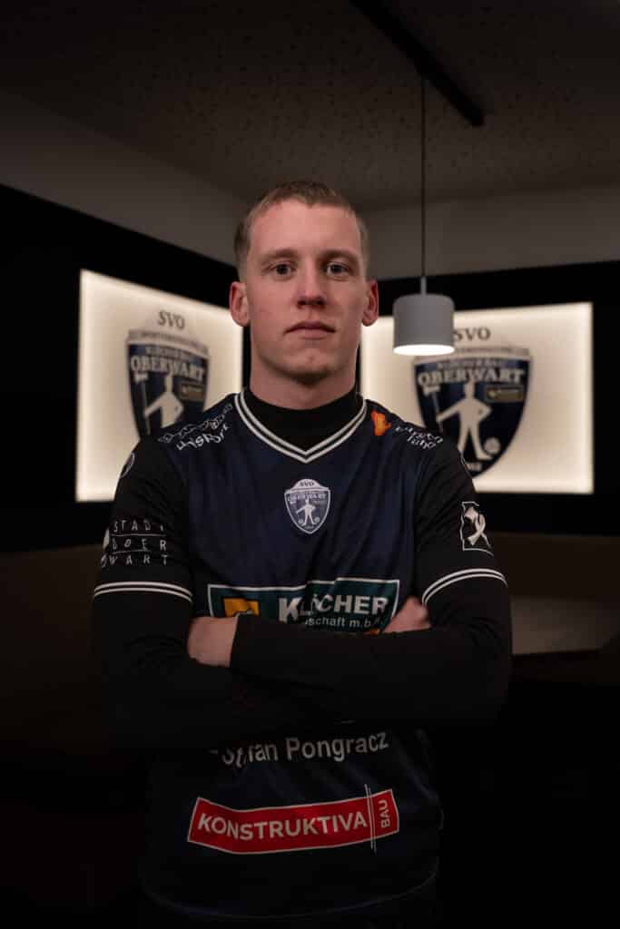 Fußballspieler im SVO Oberwart Trikot steht mit verschränkten Armen vor Vereinslogo im Hintergrund.