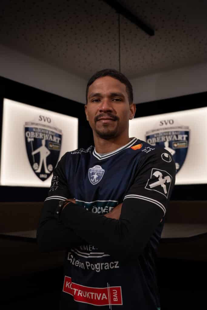 Fußballspieler in Oberwart-Trikot posiert vor Vereinslogo, Arme verschränkt, zeigt Entschlossenheit und Teamstolz.
