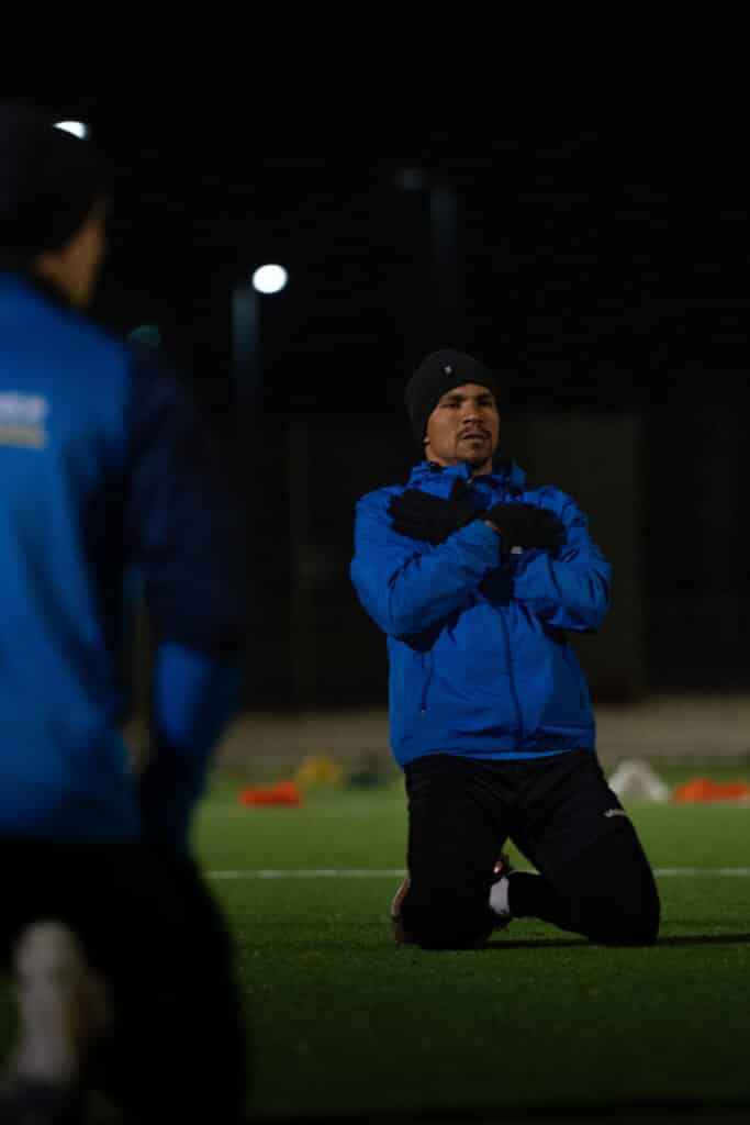 Mann trainiert im Freien bei Nacht, trägt blaue Jacke und Mütze, auf einem Fußballfeld knieend.